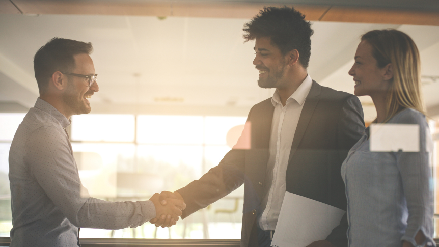 European man and woman stand side by side while the man shakes hands with another European man during a sales meeting